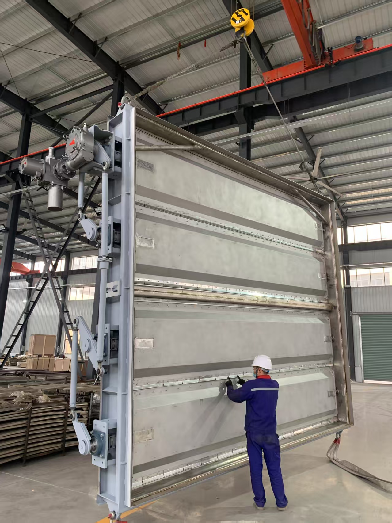 A large stainless steel electric multi louver damper is being lifted in the workshop by an overhead crane with workers conducting a factory inspection Its used for cutting off flue gas in the main desulfurization duct